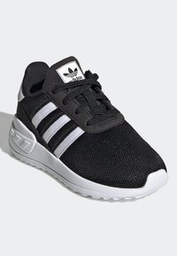 Svart idrottssko med ett mesh-övre, tre vita ränder, vadderad krage och en vit sula med struktur för bättre grepp.