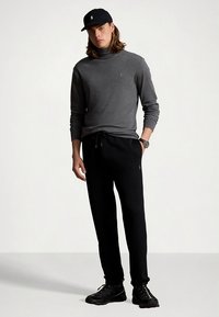 Szürke garbó pulóver, fekete, zsinórral állítható szabadidőnadrág, fekete sapka és fekete sportcipő. Egyszerű, testhezálló kialakítás, minimális díszítéssel.