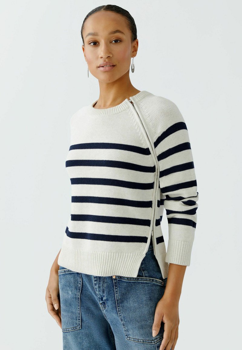 Gestreifter Strickpullover in Creme und Marineblau, mit Rundhalsausschnitt, seitlichem Reißverschluss und gerippten Bündchen und Saum. Kombiniert mit blauen Denim-Hosen.