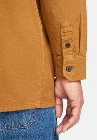 Brun texturerad skjorta med knappad manschett, med närbild av tyget och sömmarna, kombinerad med blå jeans.