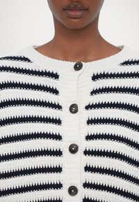 Gros plan sur une personne portant un cardigan en maille blanche avec des rayures noires horizontales et cinq boutons noirs sur le devant.