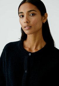 Cardigan noir, doux, avec un col côtelé et une fermeture à boutons à l'avant. Dispose de deux boutons noirs sur la patte de boutonnage pour le fermoir.