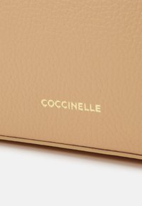 Beżowa skórzana powierzchnia o fakturze z wytłoczonym napisem "COCCINELLE" w małych złotych literach w pobliżu dolnej krawędzi.