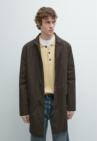 Braune, lange Jacke mit Kragen, Knopfverschluss und Seitentaschen. Wird über einem gelben Poloshirt und blauen Jeans getragen. Strukturierter Stoff.