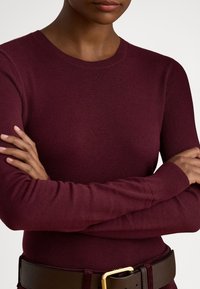 Burgundský dlhý sveter s okrúhlym výstrihom a manžetami s pružným lemovaním, nosený s hnedým koženým opaskom so zlatou prackou.