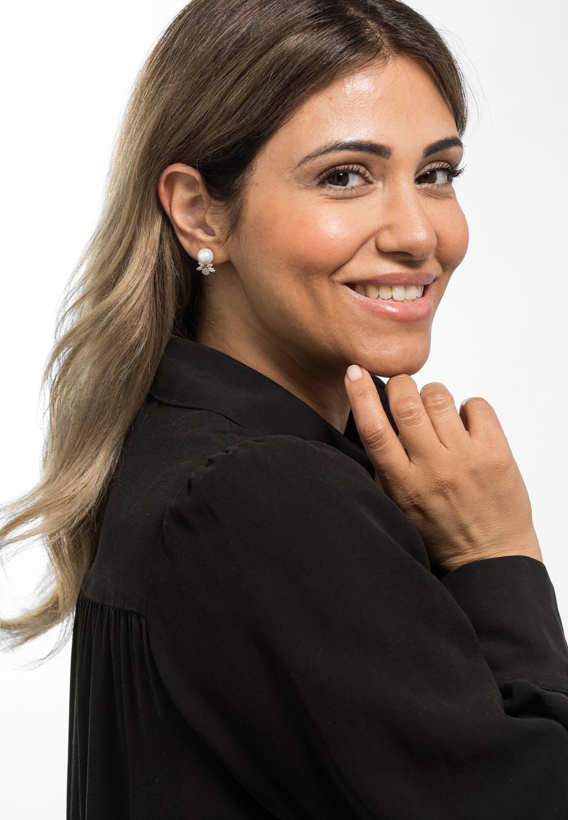 Orecchini di perle con un design floreale in oro, posizionati su una camicia nera a maniche lunghe. I capelli sono ondulati e scendono oltre le spalle.