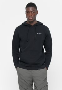 Felpa nera con cappuccio e lacci, tessuto morbido, tasca frontale e logo Columbia bianco sul lato sinistro del petto. Indossata con pantaloni cargo grigi.