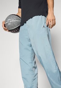Lys blå sportbukser med struktureret overflade og sort Jordan-logo, holdt med en hånd, der griber en sort og grå basketball.