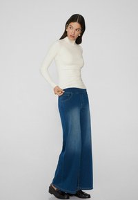 Blauwe wijd-val jeans met een vervaagde afwerking en opgestikte zakken, gecombineerd met een strakke crèmekleurige coltrui. Eenvoudige zwarte instappers maken de look compleet.
