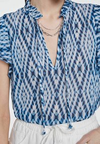 Blouse à motif bleu et blanc avec col à volants, décolleté en V profond et liens. Le tissu est léger et semi-transparent, avec un design dynamique.