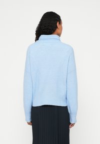 Svetlomodrý pletený sveter s vysokým golierom, nízkymi ramienkami a rebrovaným lemom. Textúra vyzerá mäkko a pohodlne. Nosený cez čiernu plisovanú sukňu.