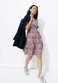 Robe à motifs paisley violets et oranges avec des manches courtes et un ourlet volanté, associée à une veste sombre et des baskets argentées.