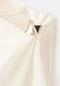 Blusa blanca con tejido suave y detalle drapeado, acentuada por un alfiler metálico y fluido en forma ondulada. Textura suave, diseño elegante.