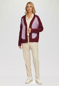 Burgundry- und lavendelfarbenes Strickcardigan mit V-Ausschnitt und Knopfverschluss, getragen über einem beigen Hemd und passenden Hosen. Weiße Schnürboots.