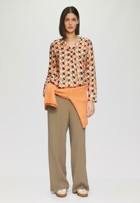 Langslavenshirt met een geometrisch patroon in oranje en bruin; gecombineerd met beige wijde broek en een oranje gebreid vest dat rond de taille is geknoopt.