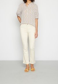 Blusa de manga corta con estampado floral en púrpura claro, con cuello de camisa abotonado. Combinada con jeans de campana crema y sandalias de tacón amarillas.