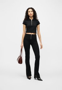 Γυναίκα με σγουρά μαλλιά που φοράει μαύρο κοντό τοπ και μαύρο παντελόνι με φερμουάρ, κρατάει καφέ τσάντα και στέκεται μπροστά σε απλό φωτεινό φόντο.