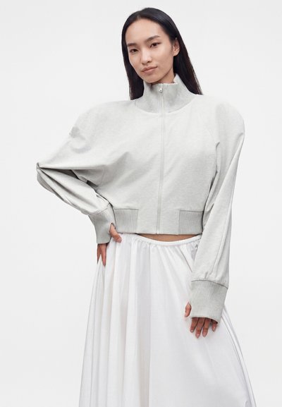 Femme aux longs cheveux foncés porte un pull gris clair zippé avec des manches exagérées et une jupe blanche fluide, posant avec la main sur la hanche.