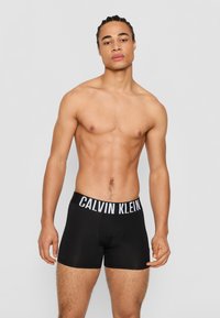 Black boxer shorts with a white "CALVIN KLEIN" logo on the elastic waistband. Smooth fabric, fitted design, mid-thigh length.