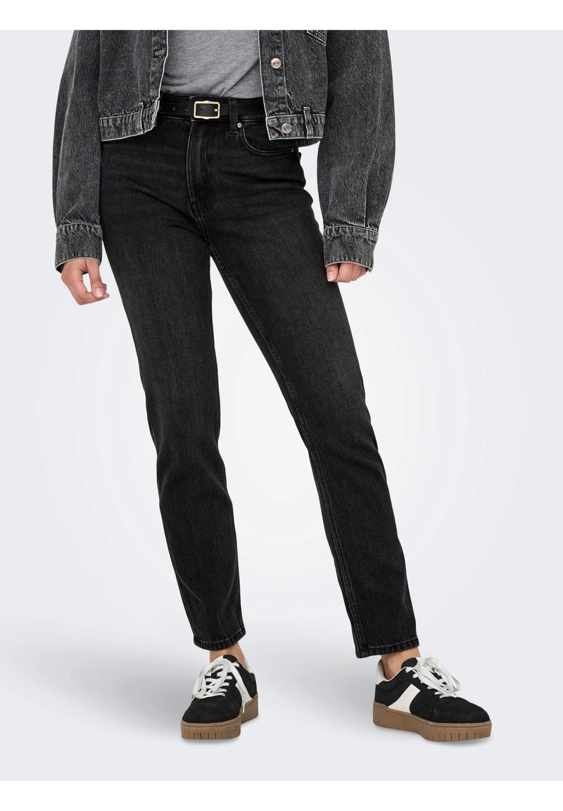 Personne portant un jean noir taille haute, une ceinture noire avec une boucle dorée, une veste en jean noire, un haut blanc et des baskets noires avec des lacets blancs et des semelles marron.
