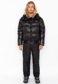 Zwarte gewatteerde jas met een glanzende afwerking en capuchon, voorzien van een ritssluiting aan de voorkant, elastische manchetten en een zwarte basislaag eronder. Zwarte cargobroek maakt de outfit compleet.
