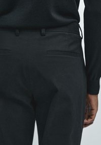 Zwarte broek van gladde stof, met twee achterzakken met gestikte randen. De tailleband heeft riemlussen en een subtiele trekkoorddetail.