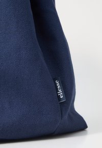 Gros plan d’un tissu bleu marine texturé avec une petite étiquette noire indiquant "ellesse" cousue dans la couture sur fond blanc.