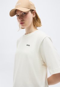 Νεαρή γυναίκα που φοράει μπεζ καπέλο BOSS και λευκό μπλουζάκι BOSS, κοιτάζοντας ελαφρώς προς τα αριστερά με ουδέτερη έκφραση, μπροστά σε απλό φόντο.