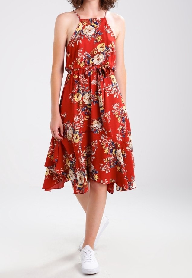 Femme portant une robe rouge sans manches à fleurs avec une ceinture à la taille et des baskets blanches, marchant sur un fond clair uni.