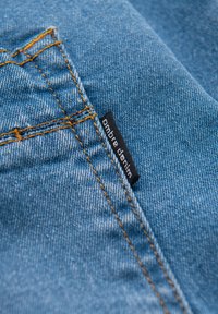 Tissu en denim bleu avec des coutures visibles en orange. Une étiquette noire portant l'inscription « ombre denim » cousue sur le tissu près de la couture.