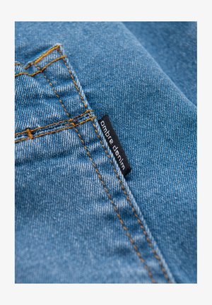 Tissu en denim bleu avec des coutures visibles en orange. Une étiquette noire portant l'inscription « ombre denim » cousue sur le tissu près de la couture.