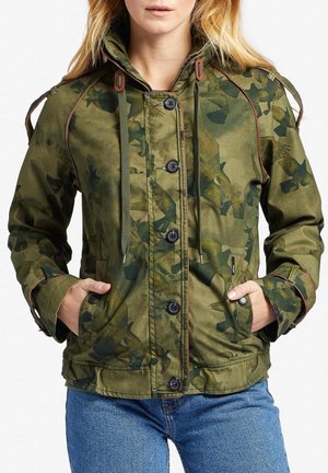 Kvinde iført grøn camouflage-mønstret jakke med knapper og snørejusterbar hætte, hænderne i lommerne, kombineret med blå jeans.