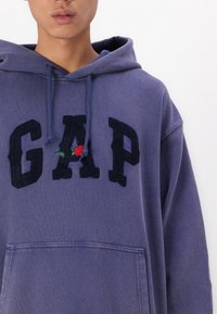 Person som bär en lila hoodie med en framficka, mörka "GAP"-bokstäver och en liten röd ros broderad på bokstaven "A."