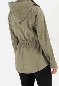 Femme portant une veste à capuche verte olive avec taille cintrée, se tenant dos à la caméra, associée à un pantalon noir.