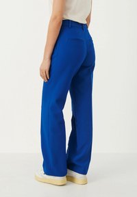 Pantaloni a gamba larga blu con una texture liscia, dotati di due tasche posteriori e una vestibilità sartoriale, abbinati a sneakers bianche.