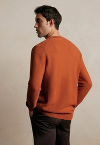 Rudawobrązowy sweter z dzianiny o okrągłym dekolcie, z teksturowanym ściągaczem i dopasowanymi mankietami, zestawiony z ciemnymi spodniami.