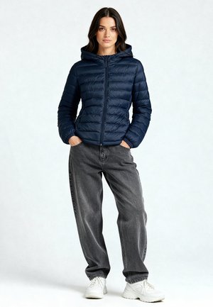 Donna in piedi con le mani nelle tasche, indossa una giacca trapuntata con cappuccio blu navy, jeans larghi grigio scuro e scarpe da ginnastica bianche su uno sfondo semplice.