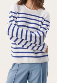 Maglione bianco con strisce orizzontali blu; texture lavorata a maglia e maniche larghe. Abbinato a pantaloni blu chiaro, che mostrano una vestibilità comoda.