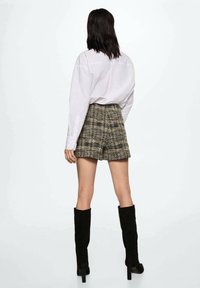 Camicia bianca con bottoni, pantaloni corti in tweed a quadri beige e neri, stivali alti fino alla coscia in suede nero, visti da dietro su uno sfondo semplice.