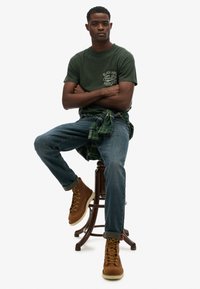 Groene katoenen t-shirt met tekst, geruite shirt om de taille, blauwe jeans en bruine veterschoenen met gele veters, zittend op een kruk.