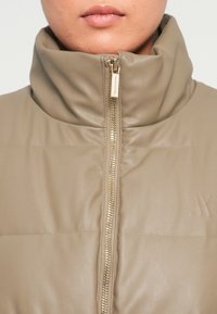 Beige gewatteerde jas met hoge kraag, rits aan de voorkant en reliëflogo-detail. Gemaakt van een gladde, strakke leerachtige stof.