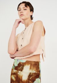Top fără mâneci de culoare piersică cu nasturi, având un design scurt. Asociat cu o fustă cu model în nuanțe de brun și verde, evidențiind texturi și culori moi.