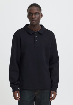 Man with dark skin wearing a long-sleeve black polo shirt and dark jeans against a plain light background.