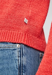 Pull rouge texturé avec un ourlet côtelé, arborant une petite étiquette de marque. Associé à un jean en denim bleu clair et à un bouton décoratif.