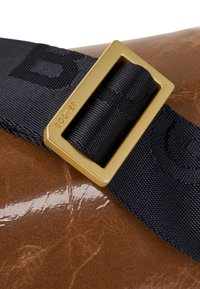 Bruine gepatineerde leren tas met een marineblauwe nylon strap. Kenmerkt zich door een gouden rechthoekige gesp, gegraveerd met "Bogner." De textuur is glanzend en glad.