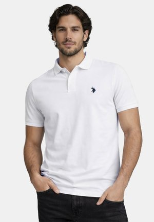 Man with dark hair wearing a white polo shirt with a small blue logo, standing with hands in black pants pockets, neutral background.