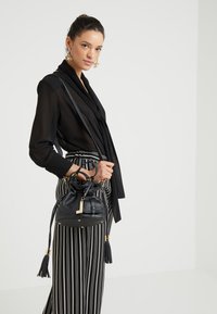 Sac seau en cuir noir avec une finition texturée, quincaillerie dorée, accents à frange et fermeture à cordon. Style de port avec une sangle.