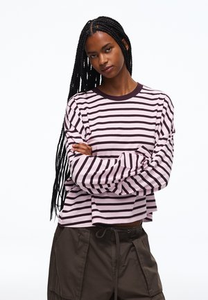 Femme avec de longues tresses portant un t-shirt à manches longues rayé rose et foncé et un pantalon cargo foncé, les bras croisés, regardant la caméra.