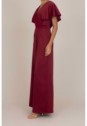 Femme portant une combinaison sans manches bordeaux profond avec jambes larges et détail d'épaule fluide rappelant une cape, associée à des talons beiges ouverts à l'avant.