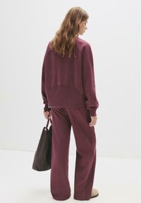 Completo sportivo bordeaux composto da un maglione a maniche lunghe con collo a girocollo e pantaloni a gamba larga. La modella tiene una borsa leggera marrone con manico intrecciato.